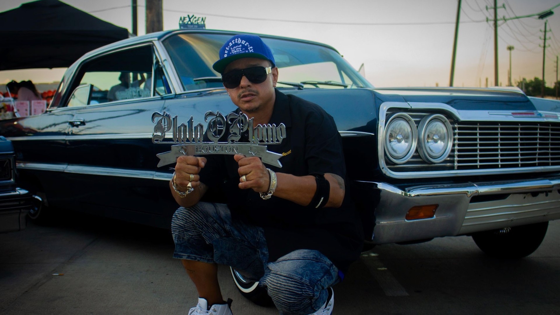 Plato o Plomo Garage, owner holding the chrome Plata O Plomo Houston plaque in front of a black 64 Impala